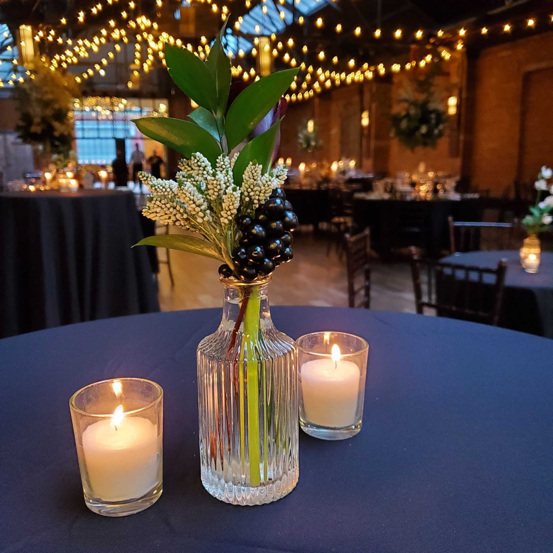 A table center piece featuring a small glass vase with greenery and dark berries, flanked by two lit candles on navy linen.
