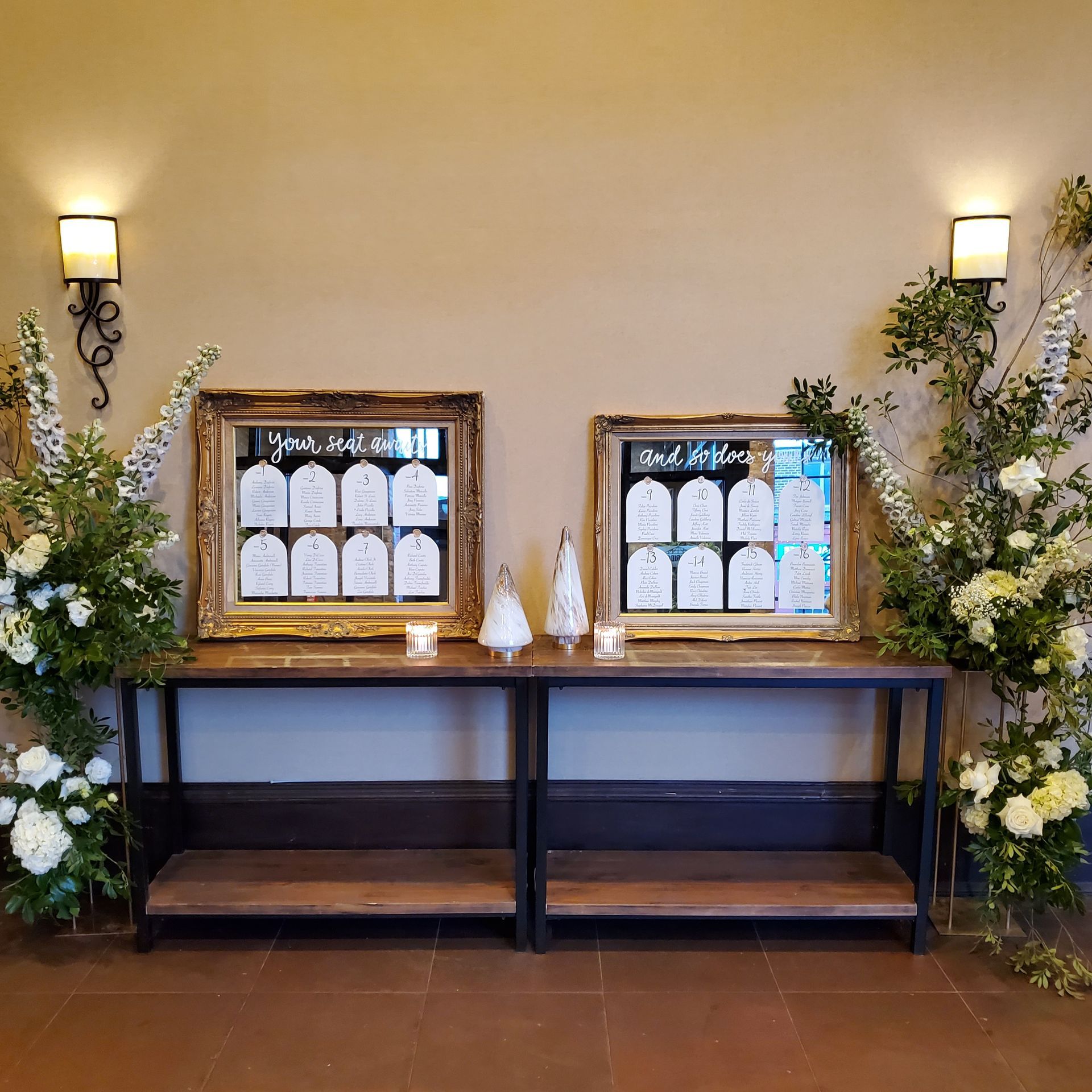 A wooden display table with two framed seating charts, candles, and large white floral arrangements at an event entrance.
