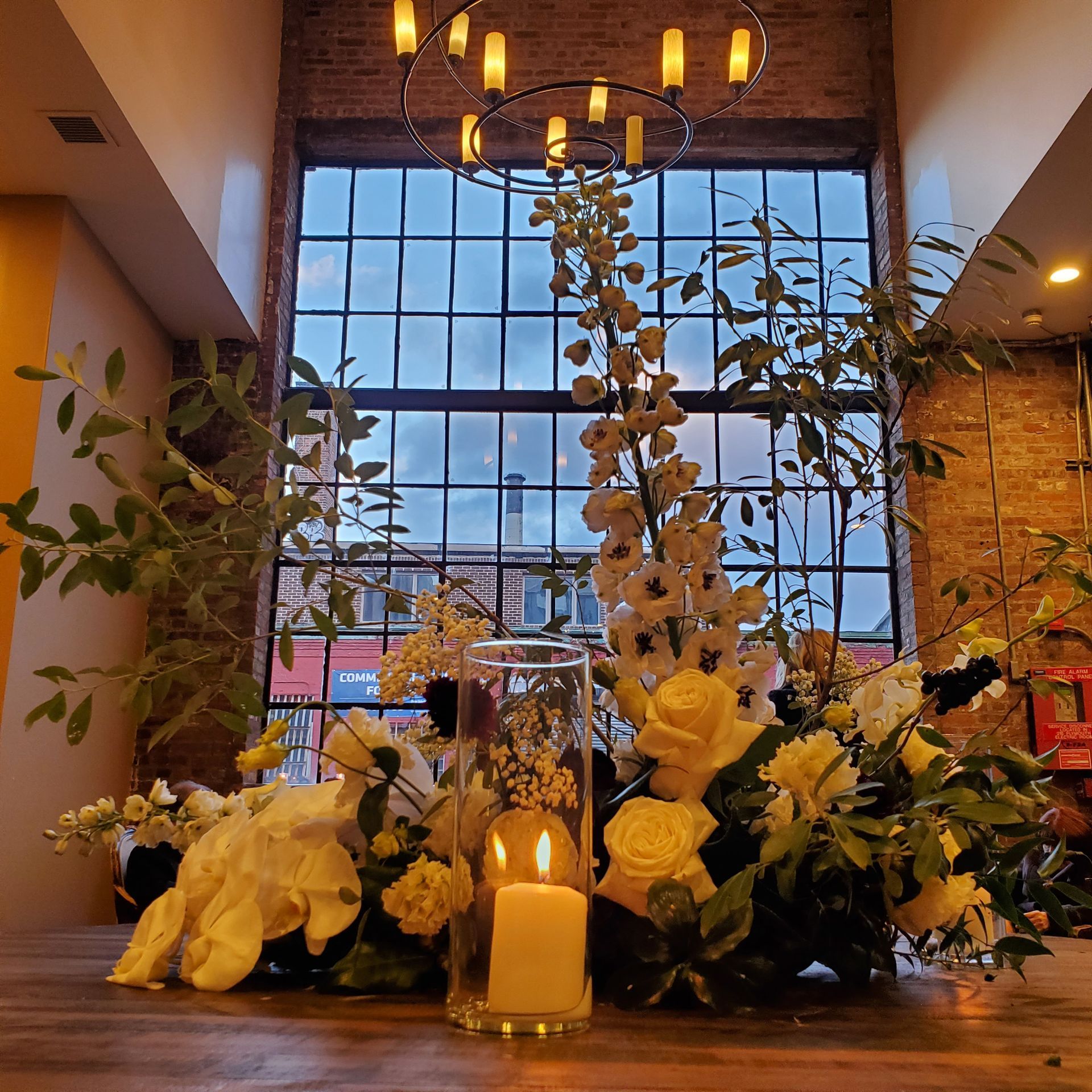 A bright, elegant floral centerpiece with yellow roses and tall white blooms surrounds a lit pillar candle on a wood table.