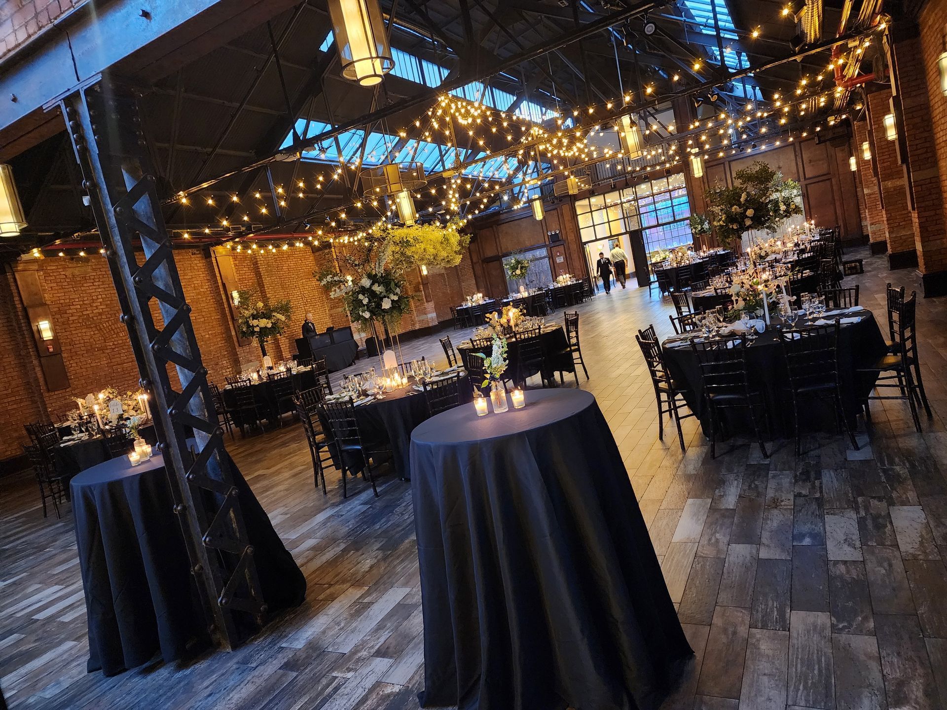 An indoor event space with dark-clothed tables, rustic brick walls, wooden floors, and glowing overhead string lights.