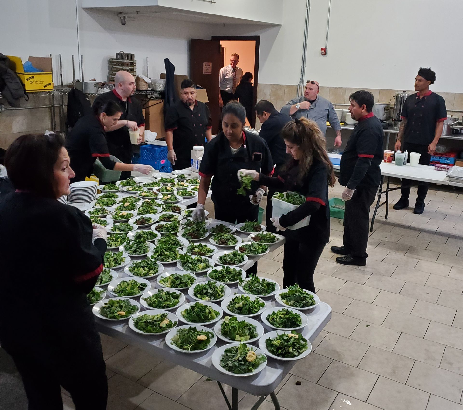 A kitchen team in black uniforms works together to prepare dozens of salad bowls on a large table.