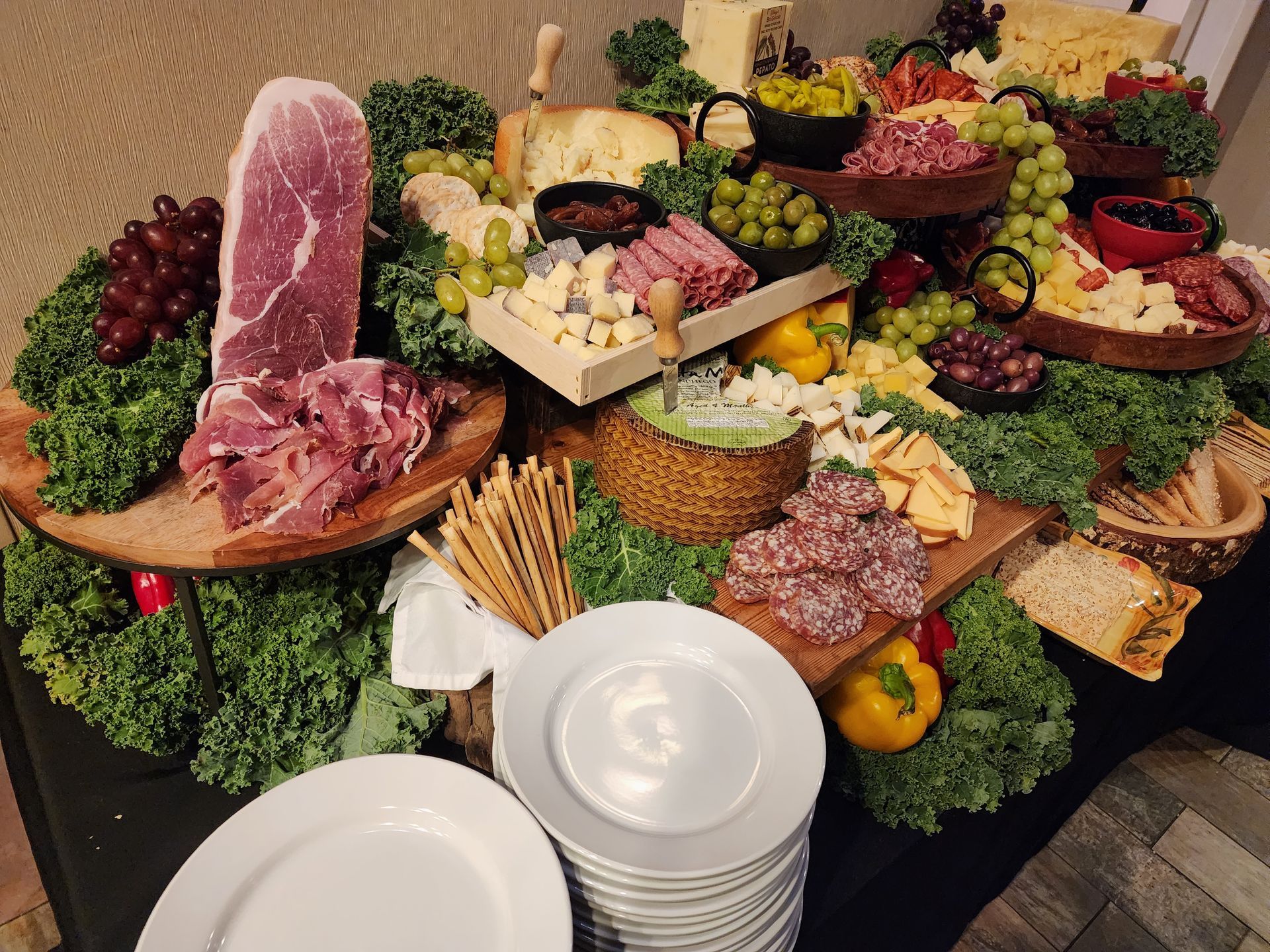 A tiered buffet display featuring assorted cured meats, cheeses, grapes, olives, breadsticks, and fresh kale garnishes.