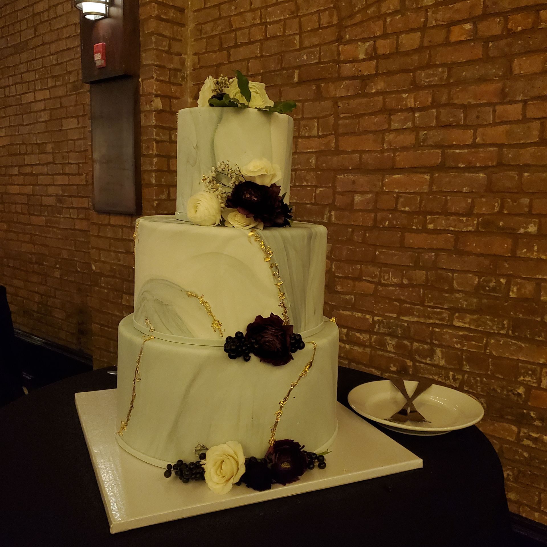 Three-tiered marble-patterned wedding cake with gold accents and dark and light floral decorations on a black table.