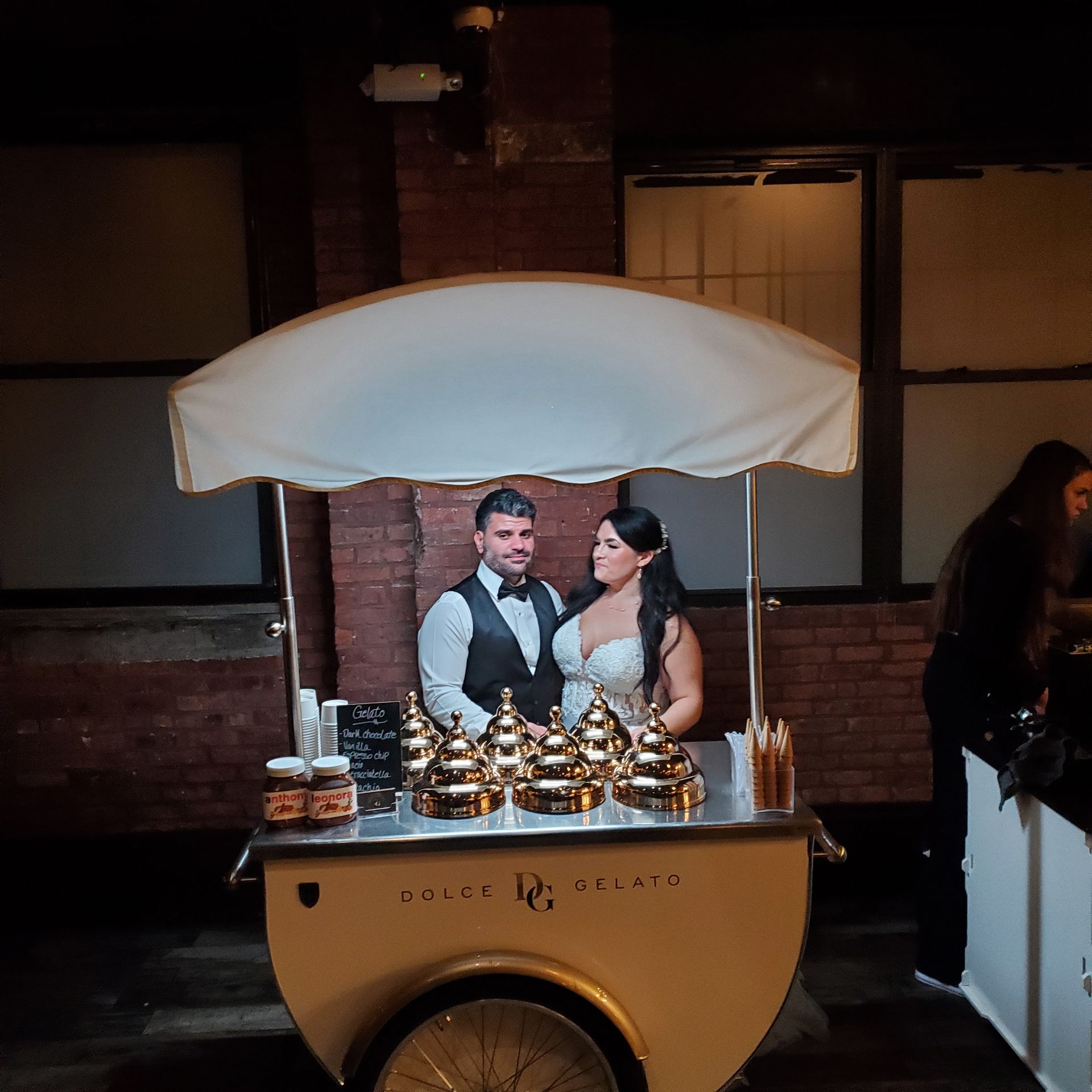 A bride and groom stand behind a cream-colored gelato cart with gold lids, illuminated against a rustic brick wall.