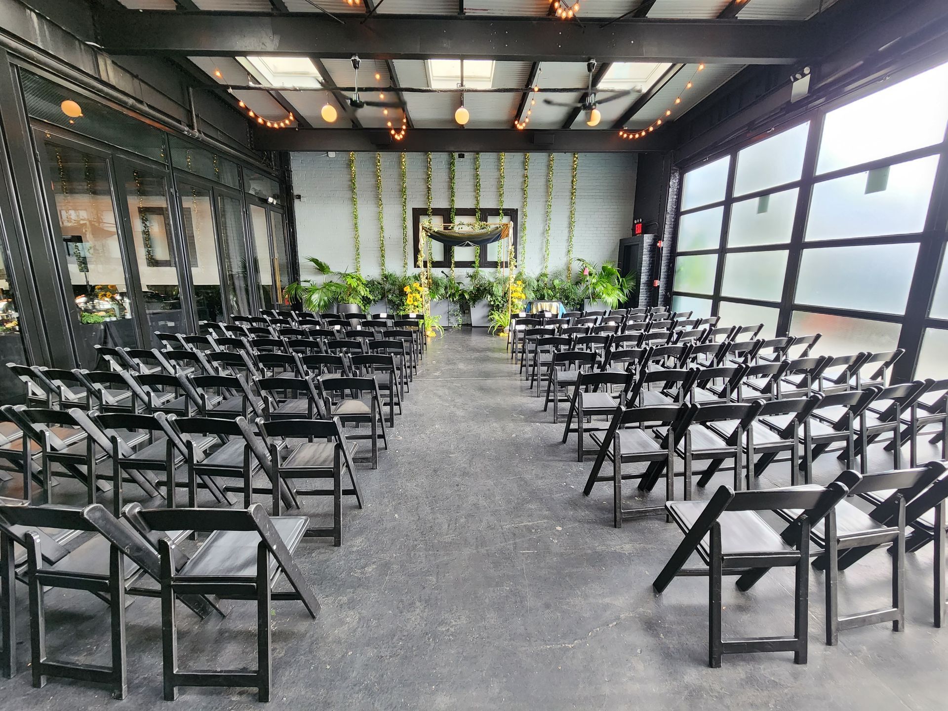 An indoor event space featuring rows of black folding chairs facing a wall decorated with greenery and a framed mirror.