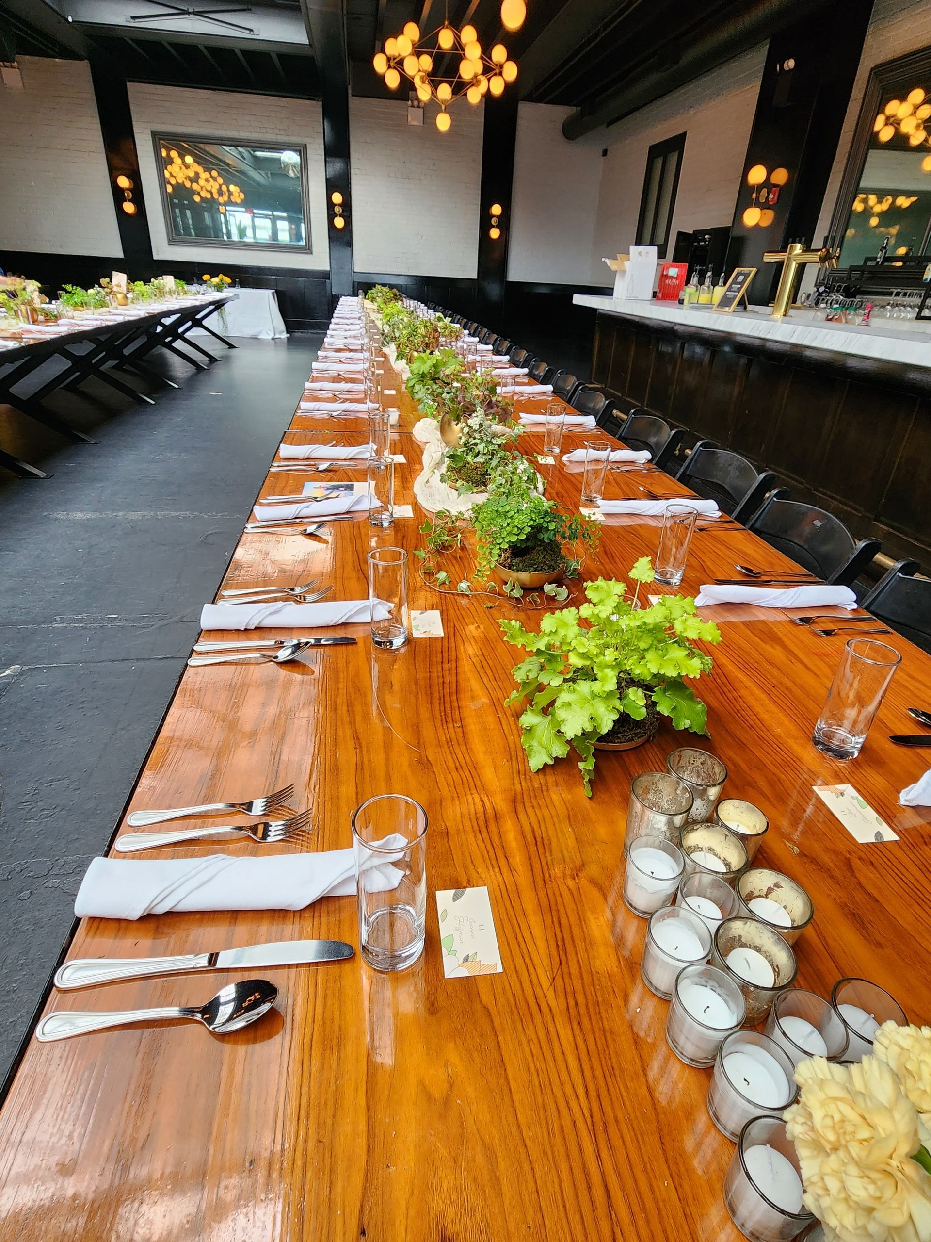 Long wooden table set for a formal dinner with place settings, floral centerpieces, and candles in a restaurant.