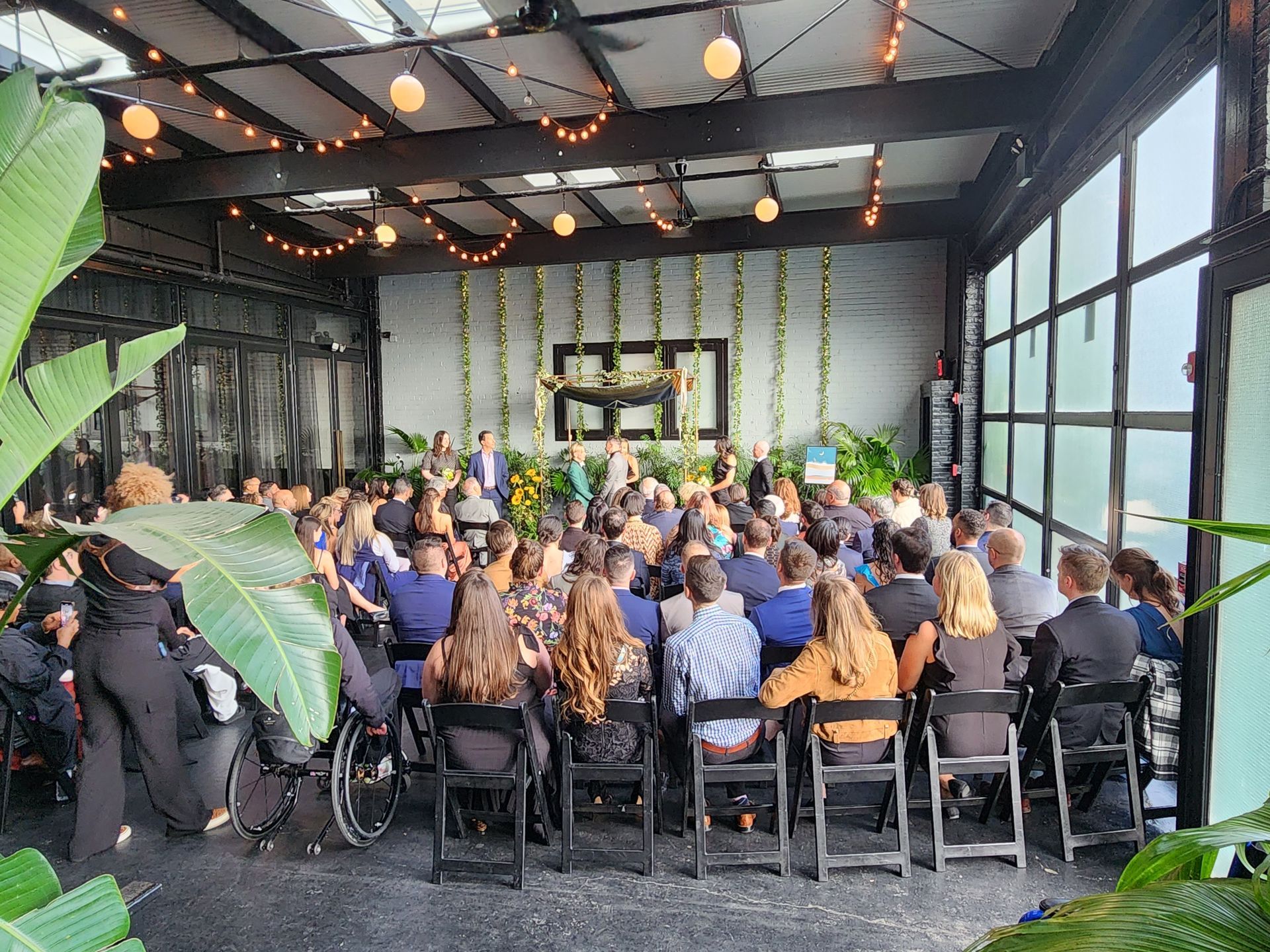 A crowd gathers for an indoor ceremony in a venue decorated with hanging lights, greenery, and a rectangular focal frame.