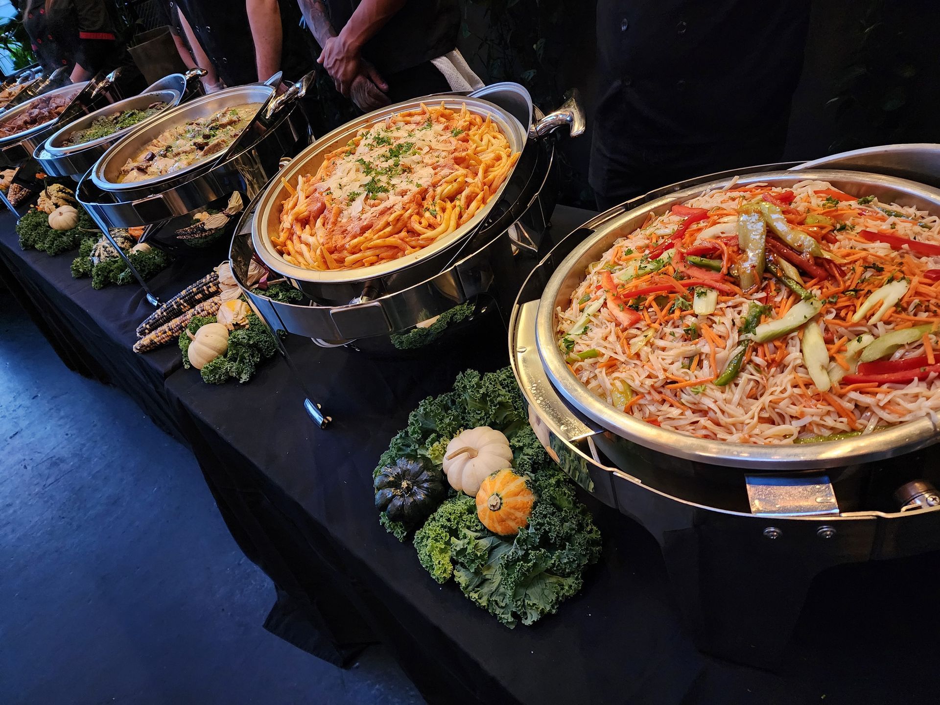 A buffet line featuring multiple chafing dishes filled with pasta and stir-fry, garnished with decorative gourds and kale.