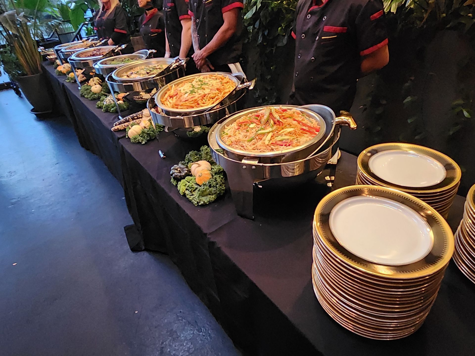 Catering staff stand behind a buffet table featuring metal food warmers and a stack of plates with gold-rimmed edges.