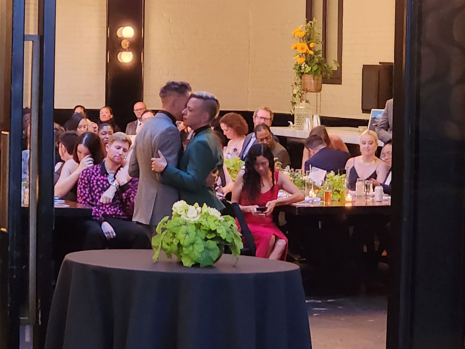 Two people dance together in the center of a crowded, dimly lit formal dining room decorated with floral centerpieces.