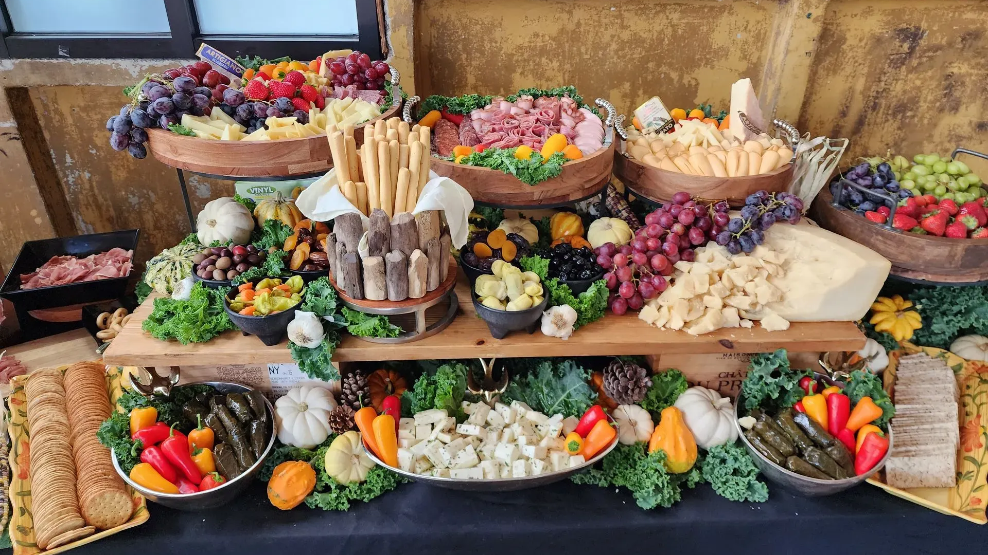A tiered buffet table loaded with assorted meats, cheeses, grapes, crackers, breadsticks, and fresh vegetables.