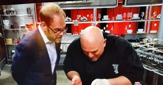 Alton Brown and a chef work in a brightly lit, red-accented industrial kitchen studio.
