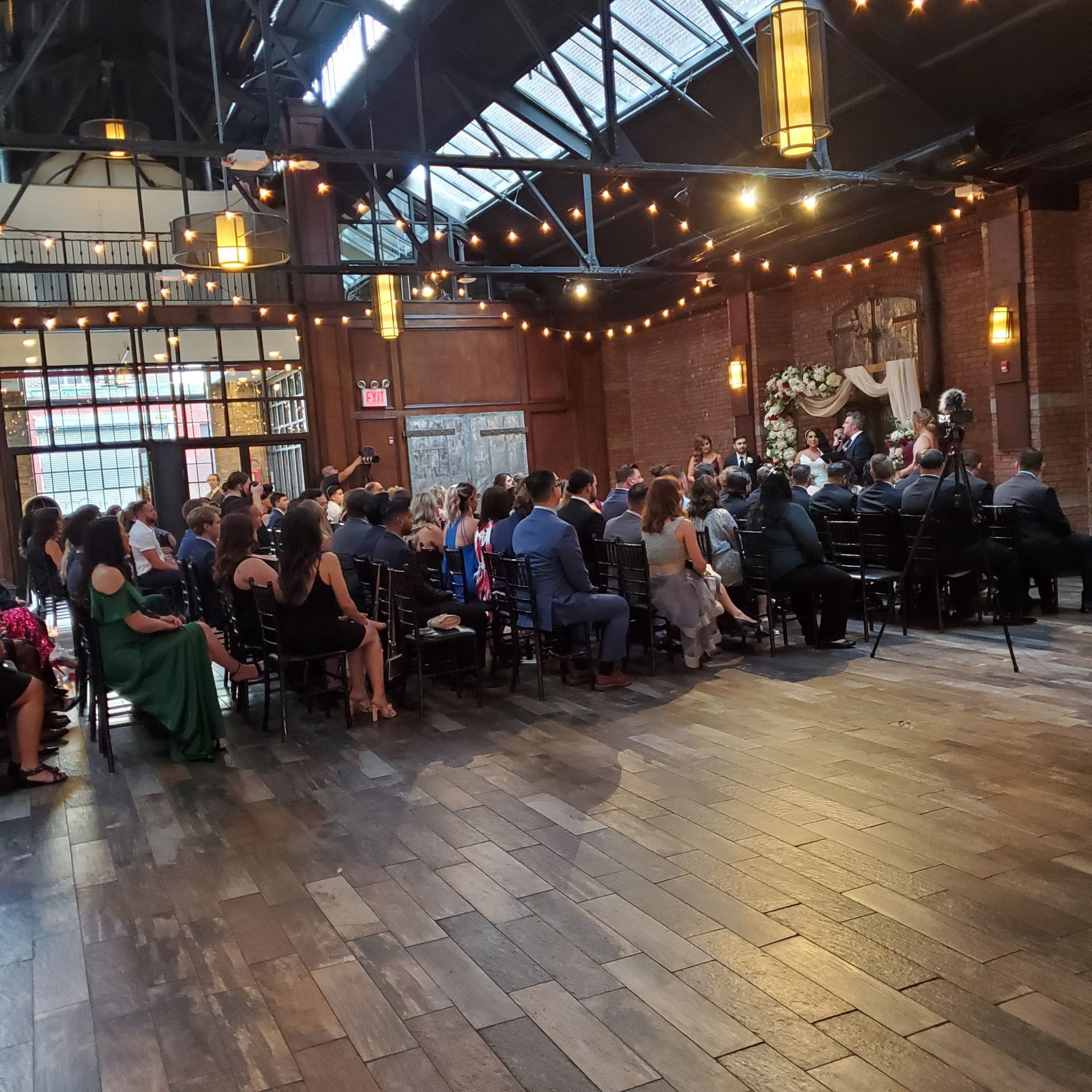 Guests sit in rows of chairs at a wedding ceremony held in a rustic, industrial venue with brick walls and string lights.