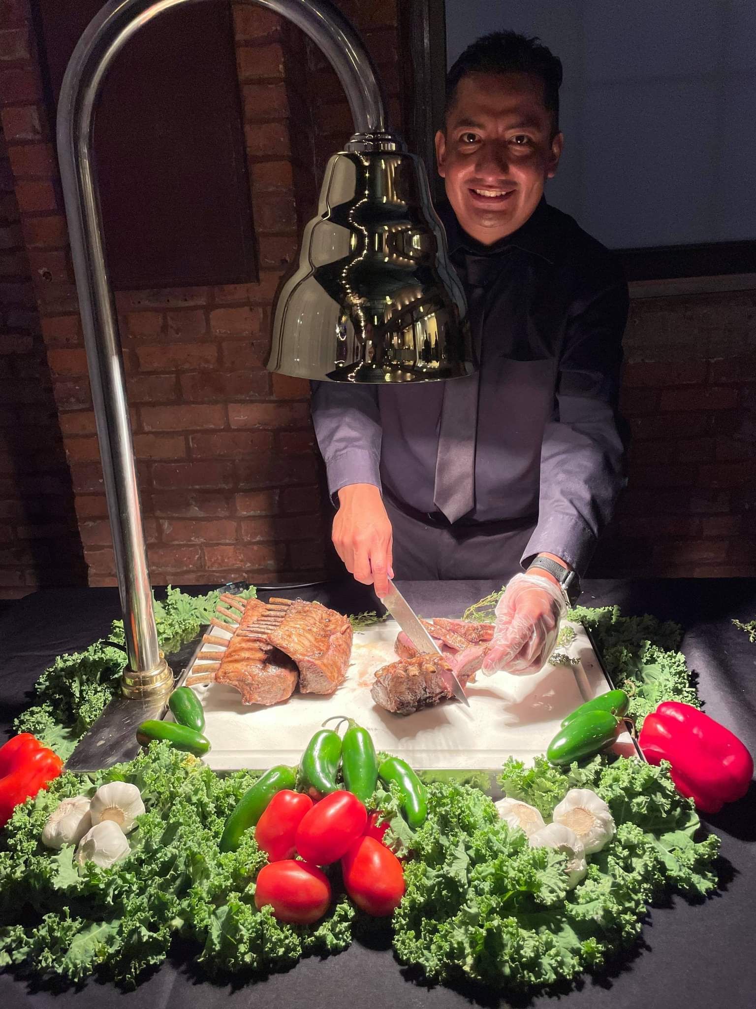 A person carves a rack of meat at a buffet station decorated with fresh lettuce, tomatoes, and peppers.