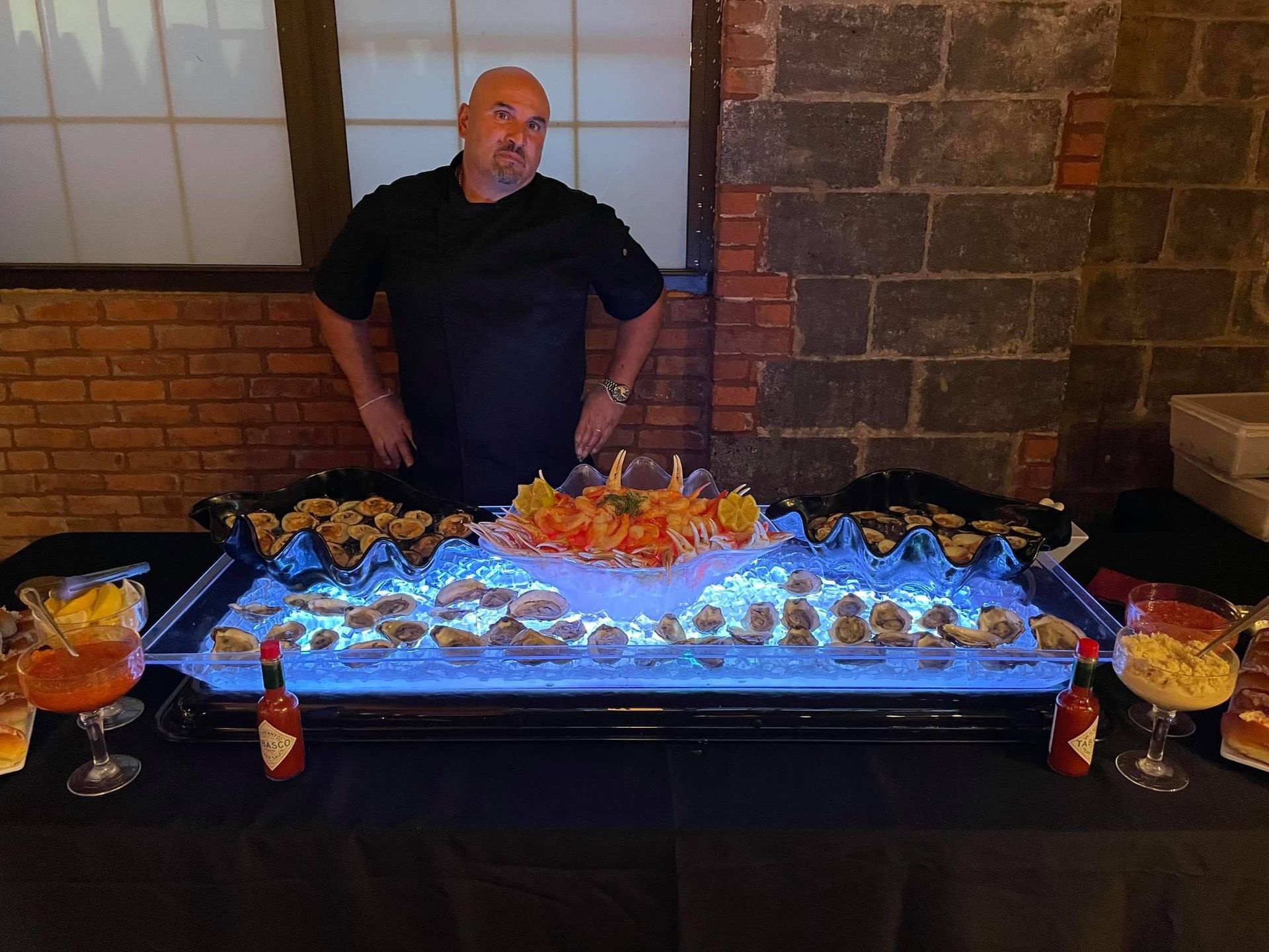 A person in a black shirt stands behind a long table serving oysters on ice with sauces, set against a brick wall background.