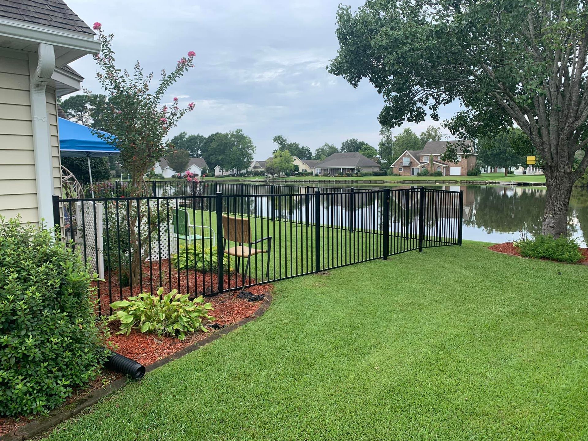 Black metal fence bordering a pond and green lawn; a house is to the left.