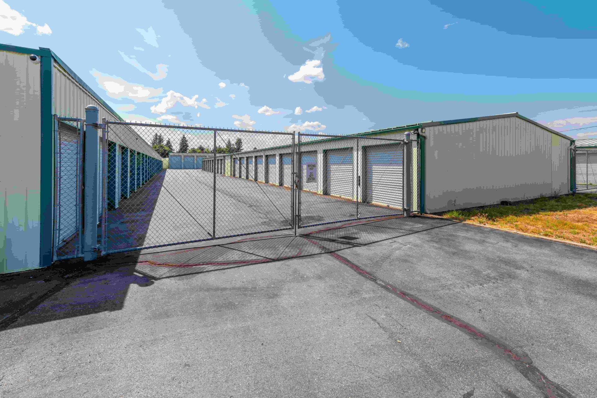 Storage units behind a chain-link fence on an asphalt lot, under a blue sky with clouds.