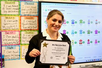 Teacher holding a certificate in a colorful classroom beside a display screen and student work posters