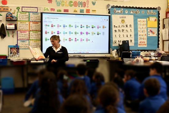 Teacher leading a classroom lesson with students seated, interactive whiteboard and colorful posters visible