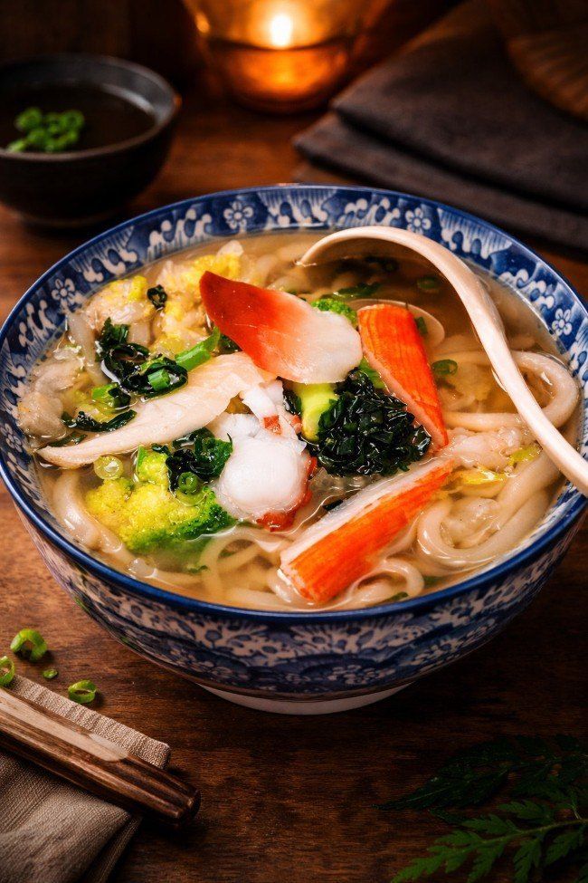 Blue bowl of noodle soup with crab sticks, shrimp, greens, and chopsticks on a wooden table