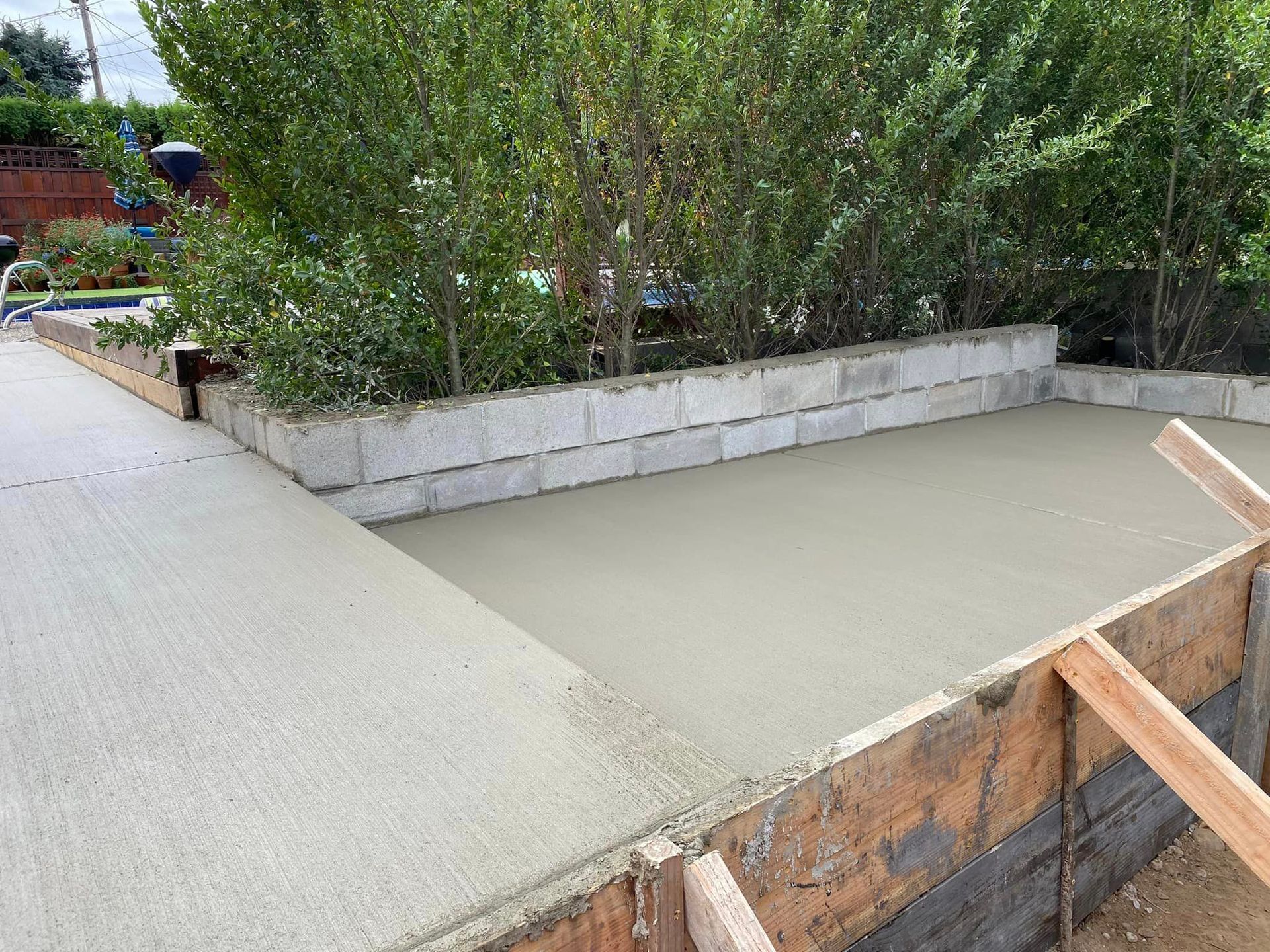 Newly poured concrete sidewalk and rectangular slab next to a low cinder block garden wall and green bushes.