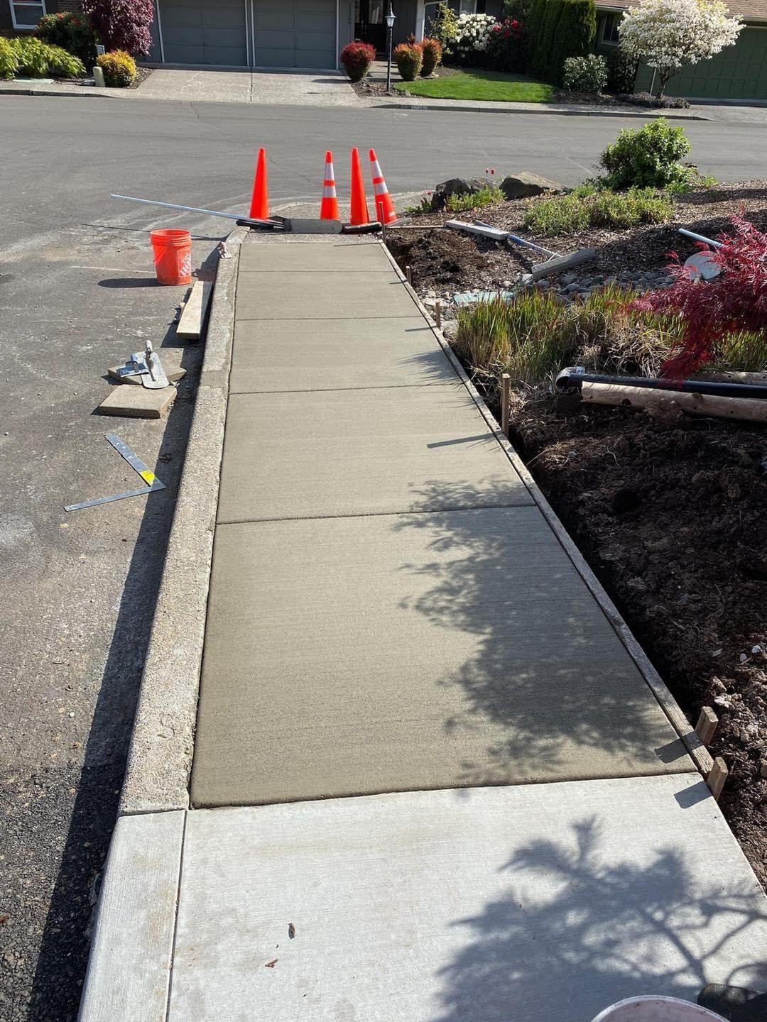 A newly poured concrete sidewalk segment under construction, cordoned off by four orange traffic cones.