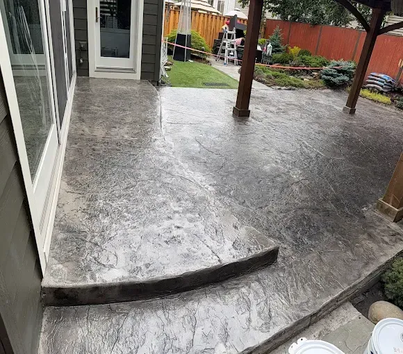 A gray stamped concrete patio with a single step leading to a house door, featuring a wooden pergola post and backyard.