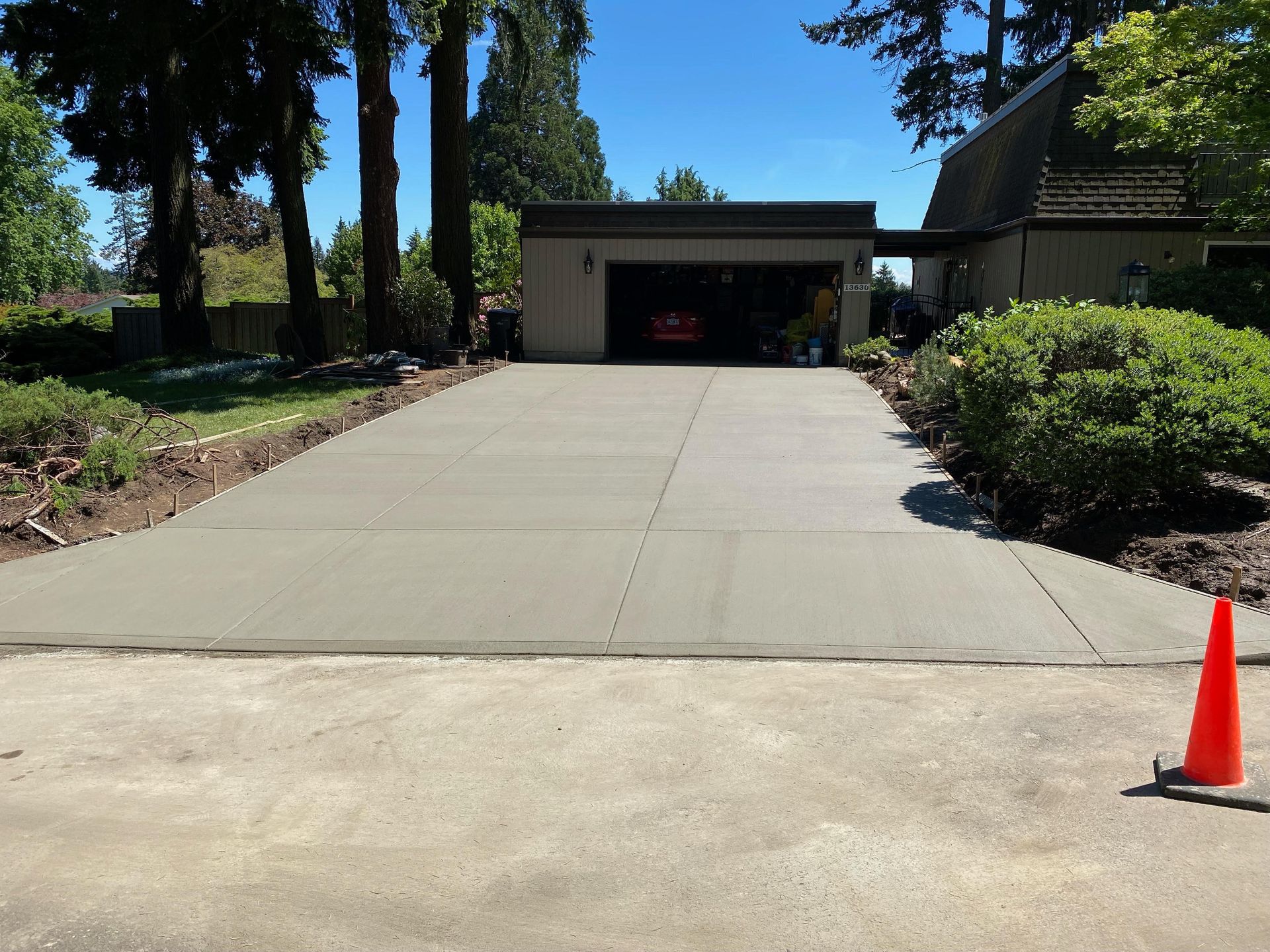 A freshly poured, smooth gray concrete driveway leads to a suburban garage on a sunny day, with a small orange cone nearby.