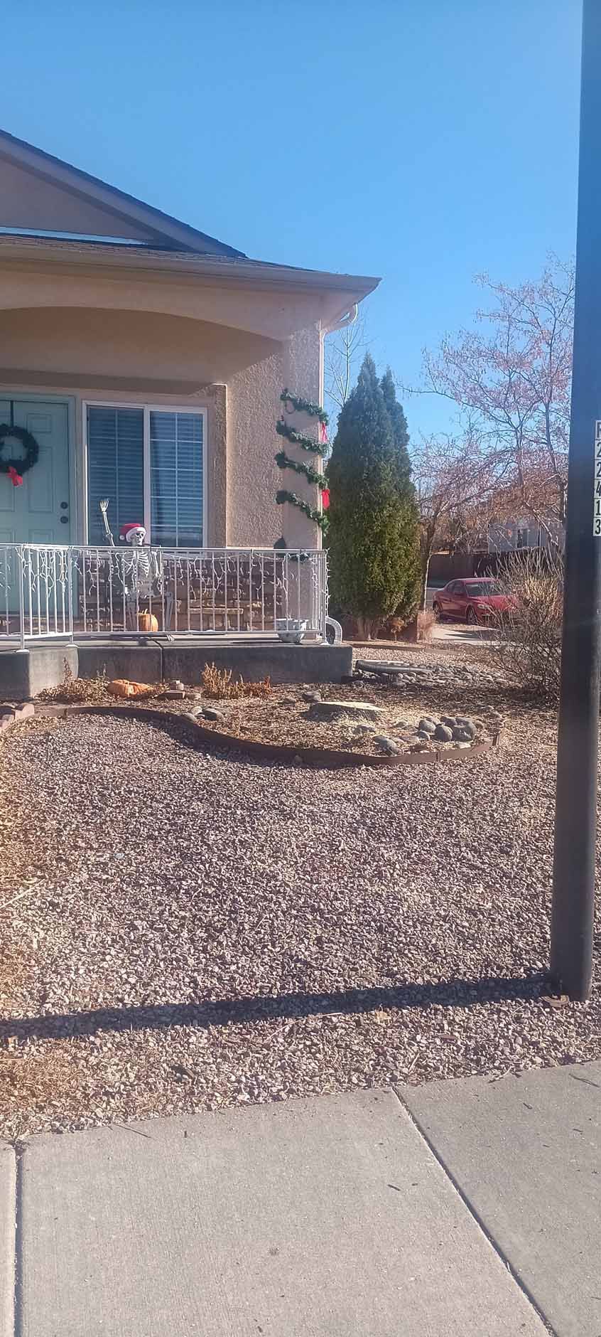 A residential house exterior with a porch, a decorated pillar, a front yard with gravel, and a sidewalk under a blue sky.
