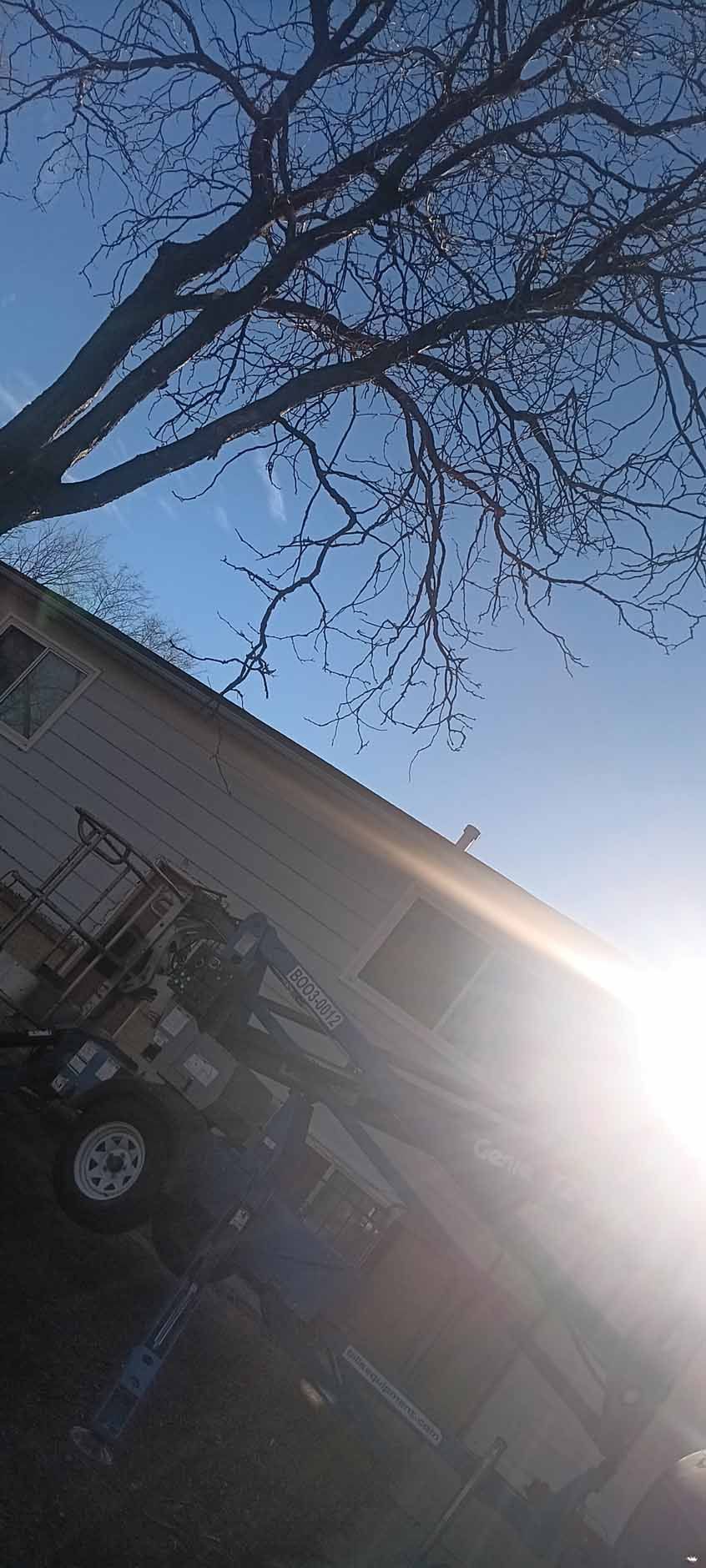 A bright, sun-drenched view of a two-story house exterior under a clear blue sky, partially shaded by bare tree branches.