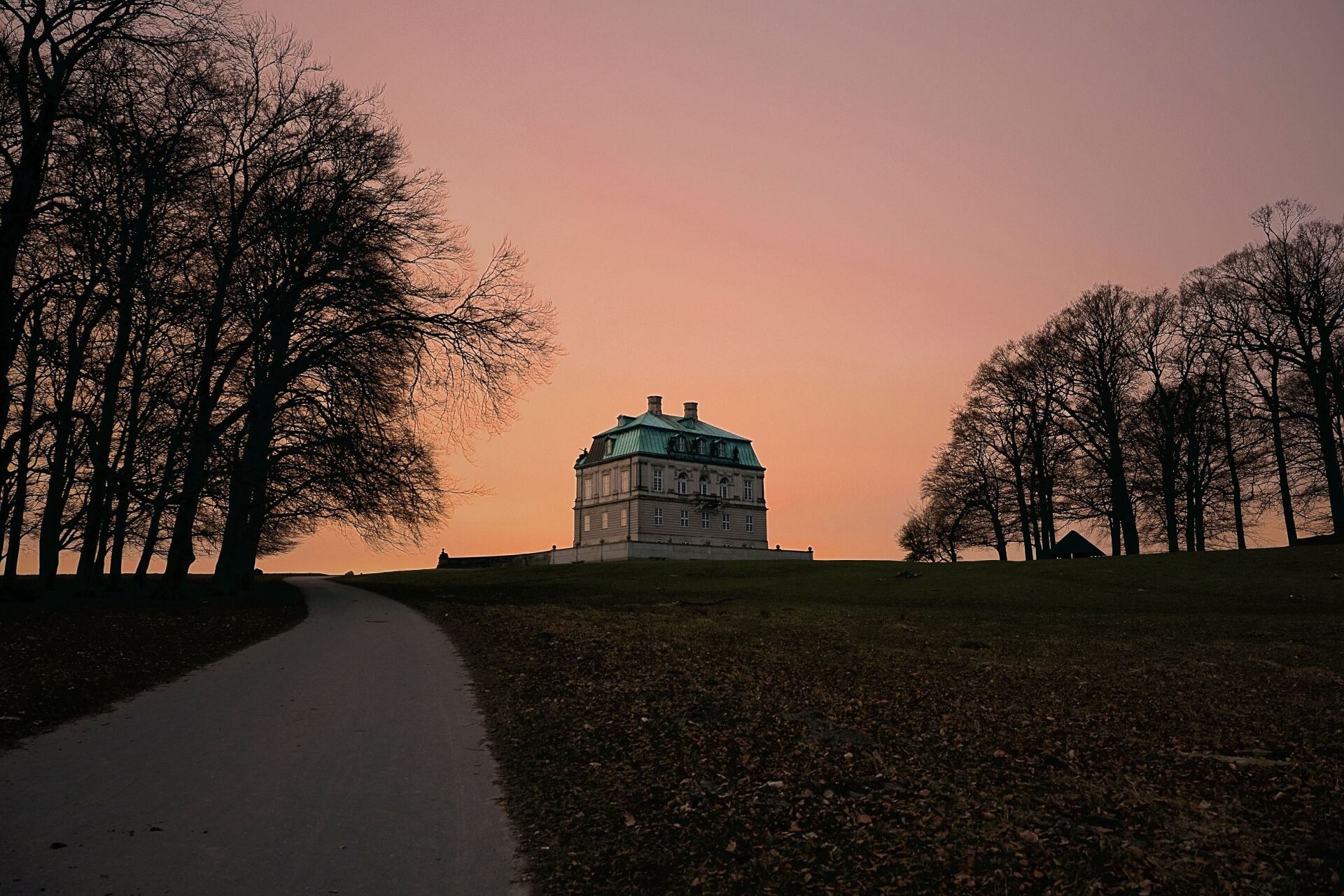 Et stort hus ligger på toppen af ​​en bakke omgivet af træer ved solnedgang.