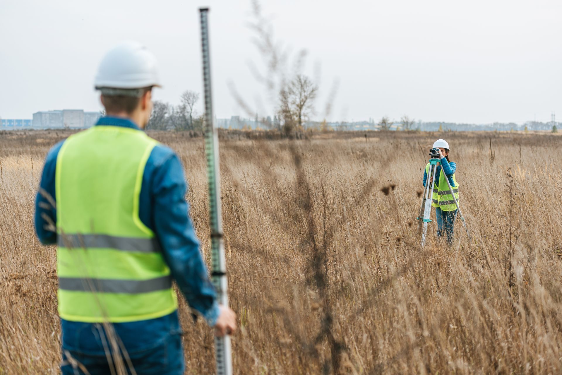 Two land surveyors in safety vests using a total station and survey rod in a grassy field