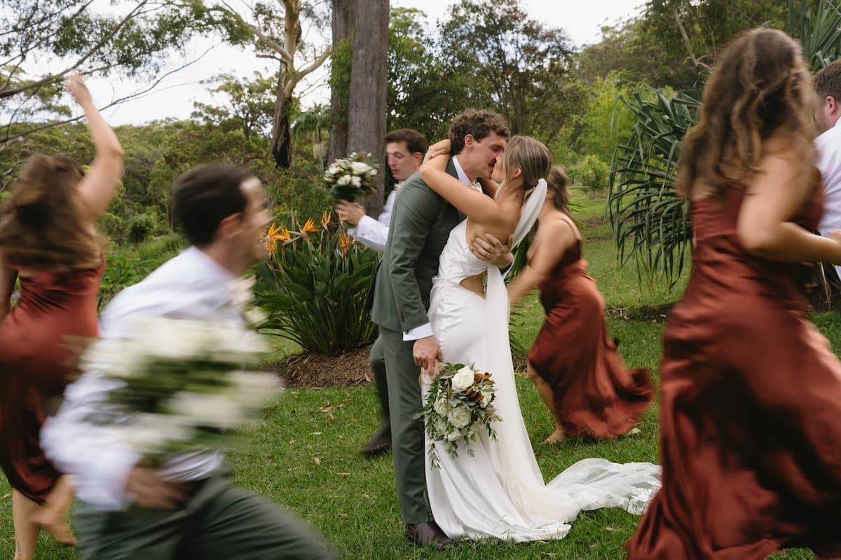 Wedding Couple Kissing Outdoors as Guests Run Past in a Grassy Garden โ Seed + Salt Photography In Smithtown, NSW