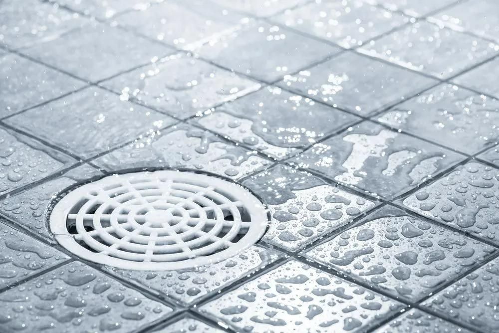A Drain on A Tiled Floor with Water Drops on It — Gillespie Plumbing in Grafton, NSW