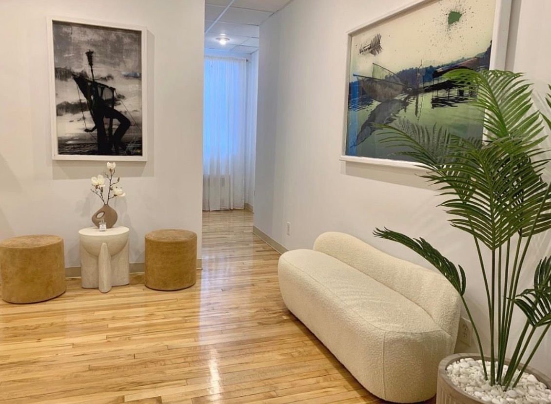 Bright minimalist living room with abstract art, tan stools, white chair, cream sofa, and potted palm.