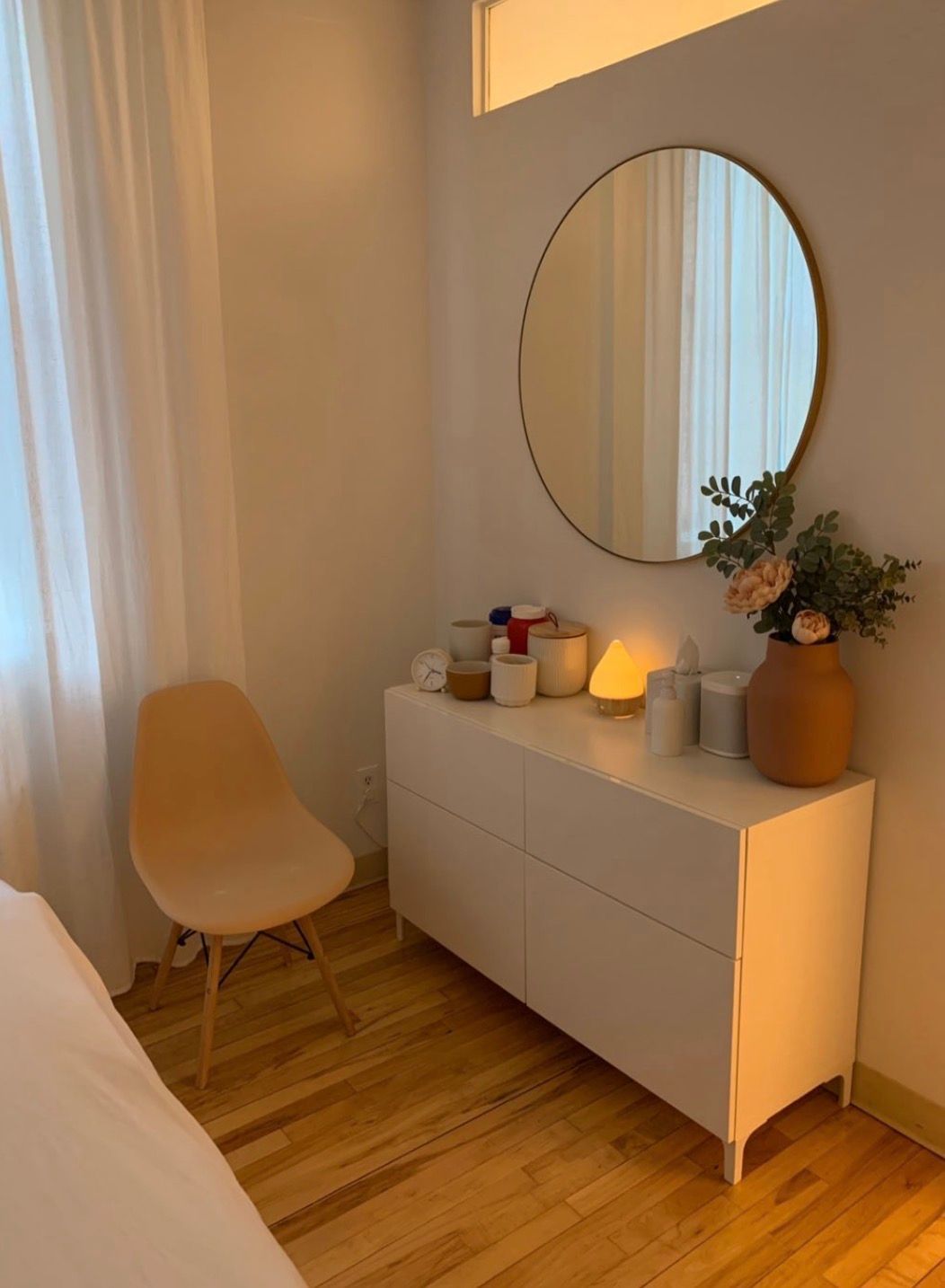 Minimal bedroom corner with cream chair, white dresser, round mirror, and vase of flowers by a window