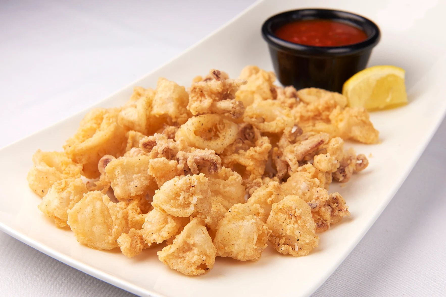 Point Judith Calamari Fritti A white plate topped with fried calamari and a small bowl of sauce.
