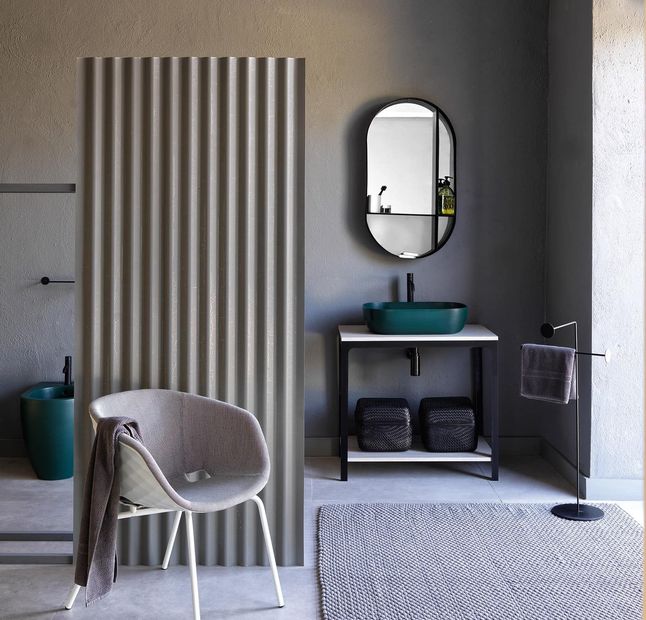 Bagno con pareti grigie, lavandino verde, divisorio ondulato, sedia e specchio ovale.