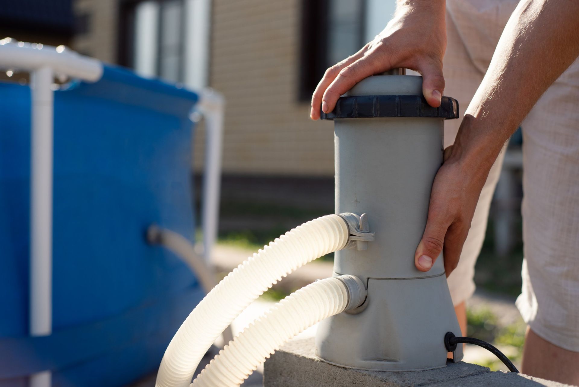 Person placing a gray pool filter next to a blue above-ground pool; outdoors.