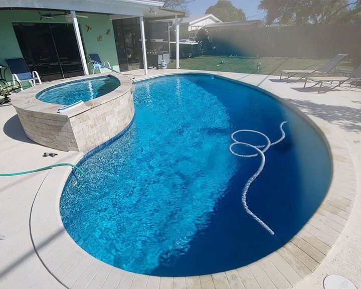 A blue-tiled swimming pool and spa surrounded by a white patio in a backyard setting. A hose is in the pool.