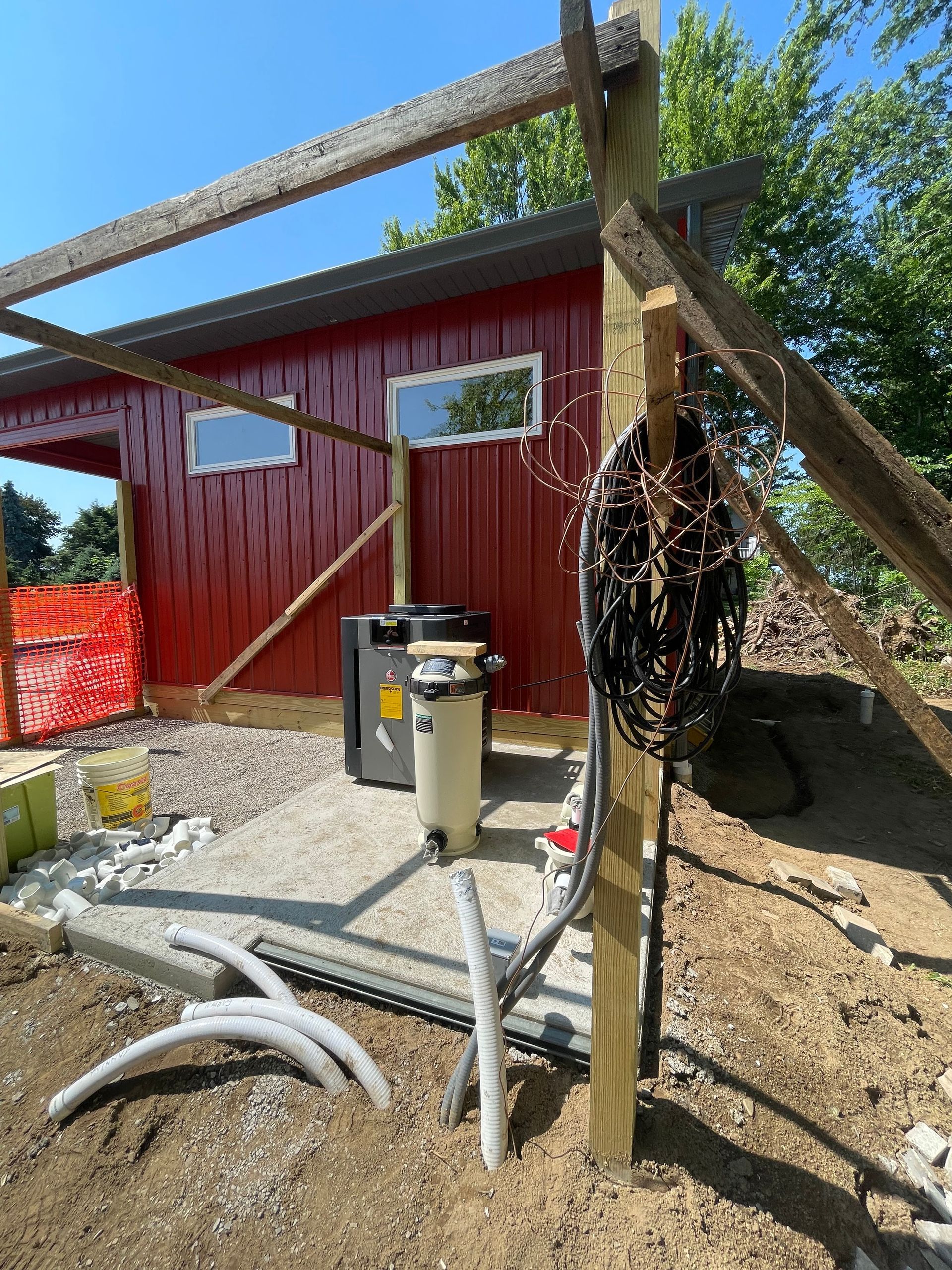 Pool equipment next to a red shed with gray gravel. Dark filter, pipes, and electrical cables present.