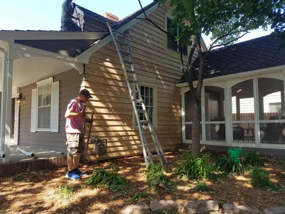 Worker on ladder repairing roof siding beside a house with a damaged roof section