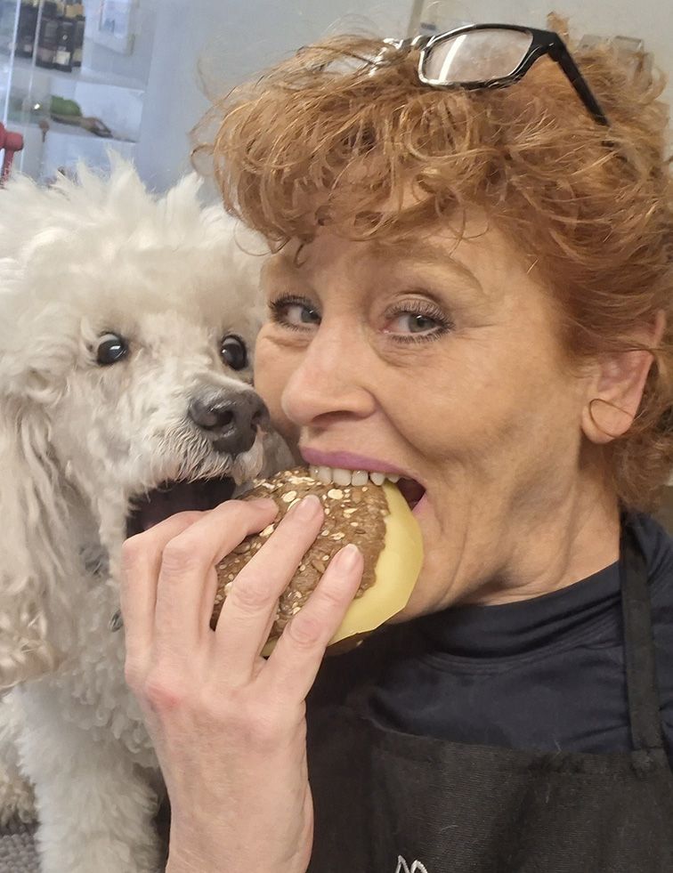 Een vrouw eet gebak met een witte hond naast haar. De hond kijkt naar het gebakje, dat dicht bij de mond van de vrouw wordt gehouden.