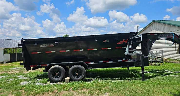 Black dump trailer with dual axles parked on grass beside a house under a partly cloudy sky