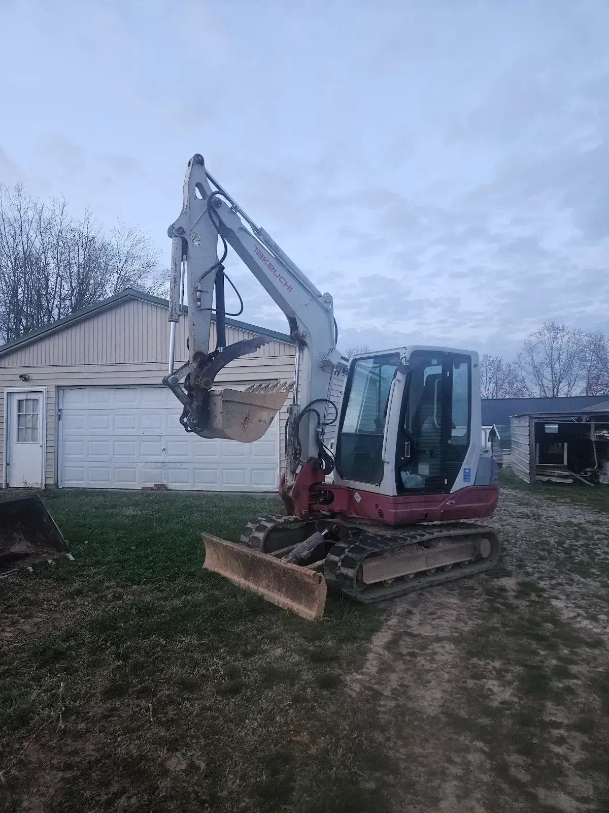 Red mini excavator with raised bucket parked on a dirt path beside a house and garage.