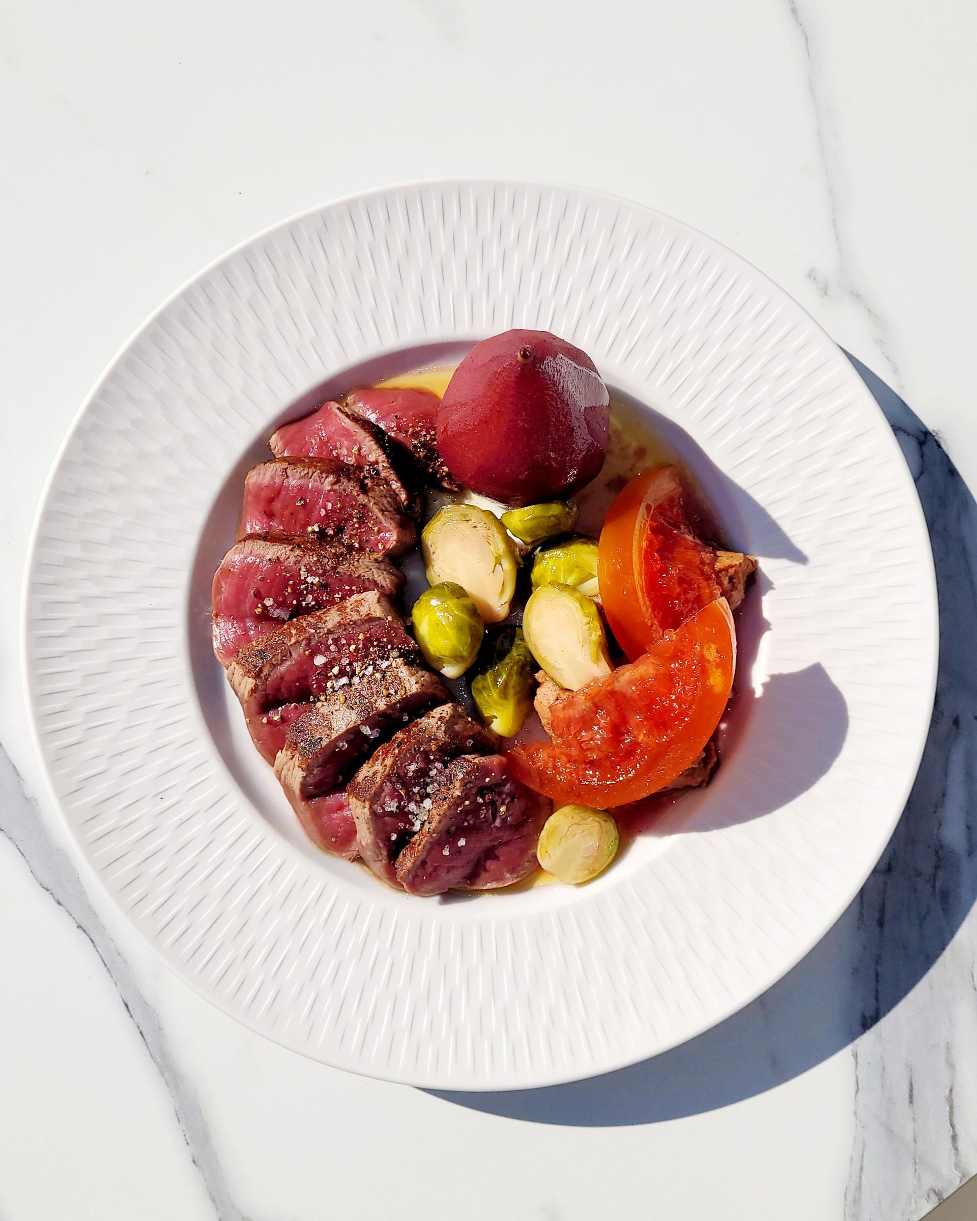 Thon saisi en tranches, accompagné de choux de Bruxelles, de tomate et d'une poire pochée, servi sur une assiette blanche.
