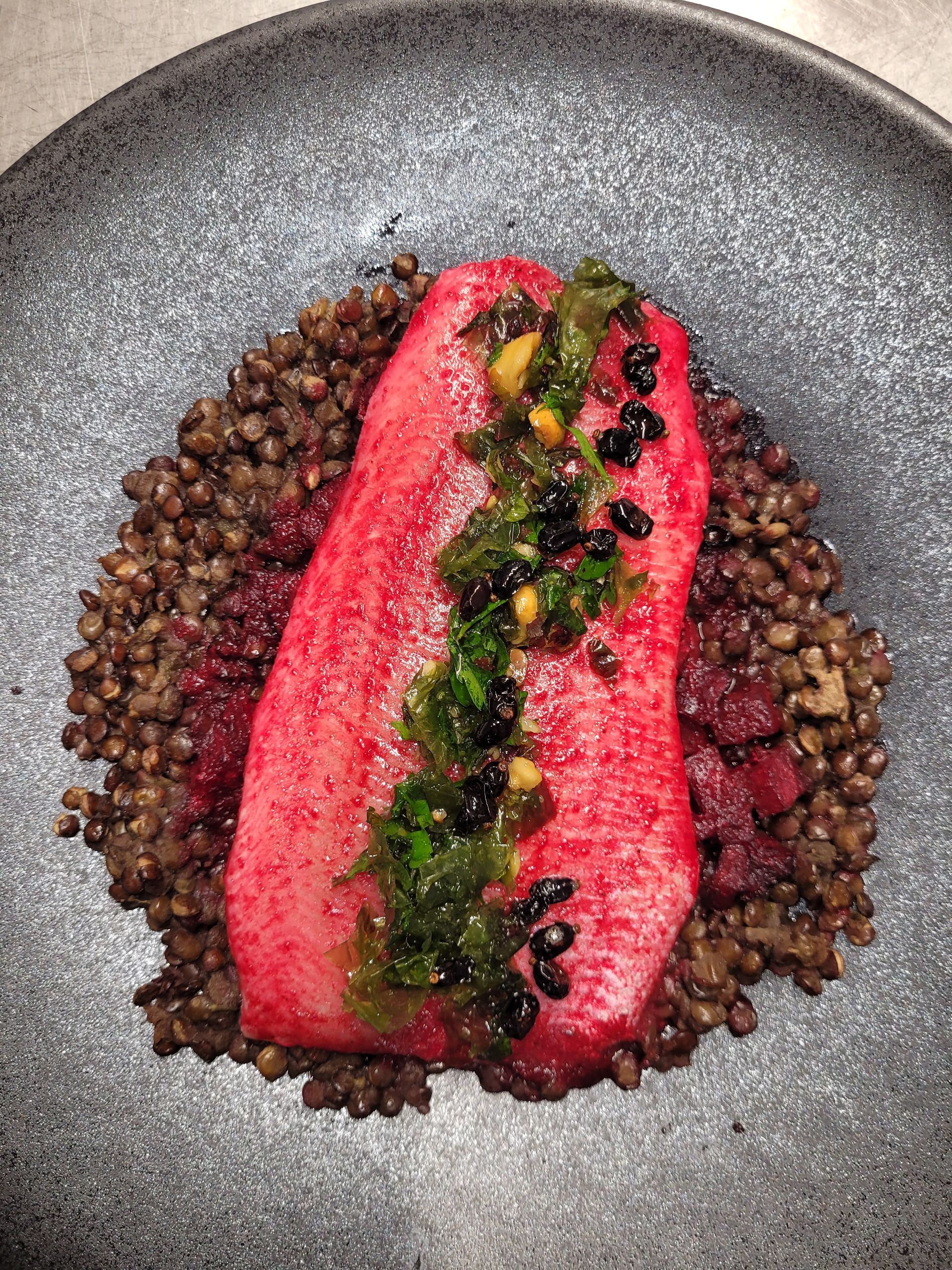 Plat aux teintes rosées sur une assiette mouchetée : base de lentilles garnie de protéines roses, d’un filet d’herbes vertes et de mûres noires.