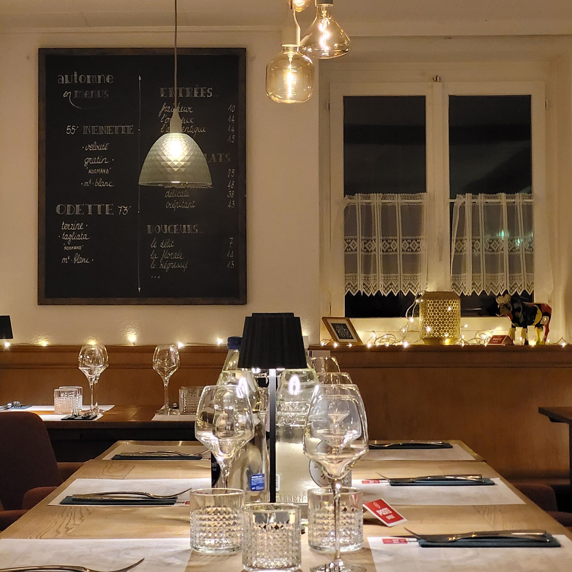 Intérieur d'un restaurant avec tables dressées pour le dîner, menu sur tableau noir, guirlandes lumineuses et fenêtre ornée de rideaux en dentelle.