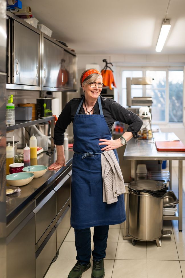 Femme en veste de chef, bras croisés, regardant sur le côté, devant un mur blanc.