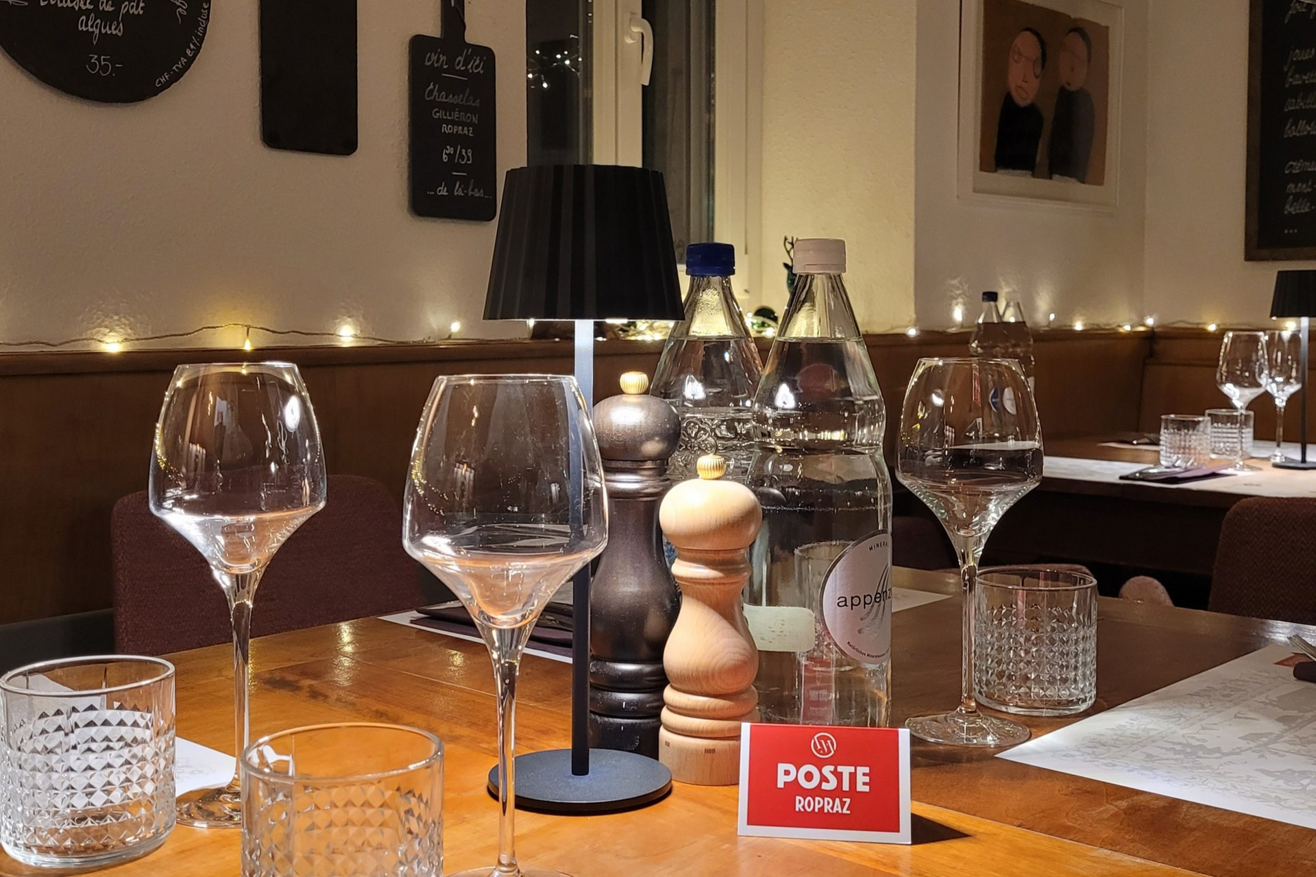 La table est dressée pour le repas : verres à vin, bouteilles d’eau, salières et poivrières, et une petite lampe.